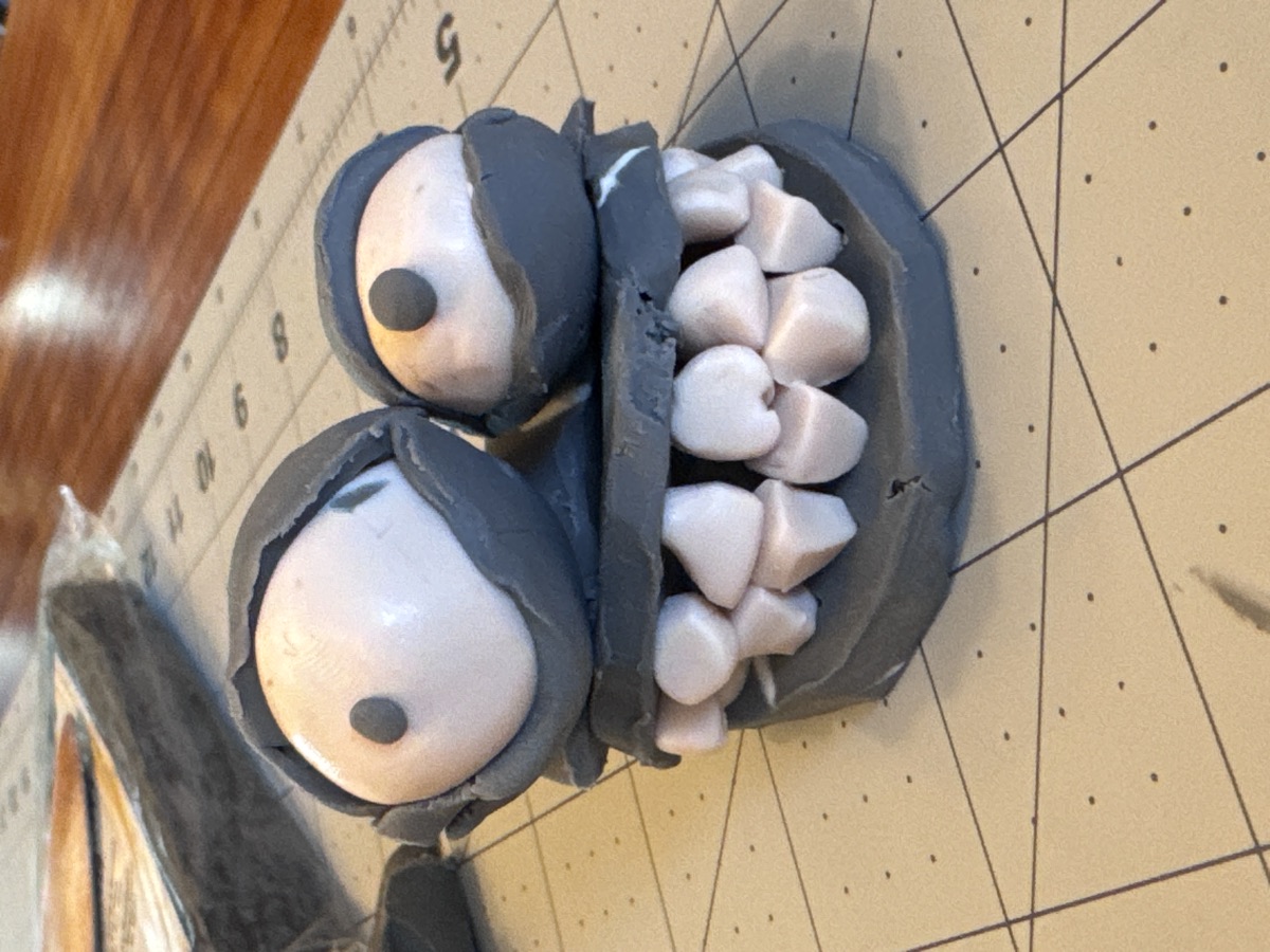 A clay monster face with googly eyes and sculpted teeth on a cutting mat